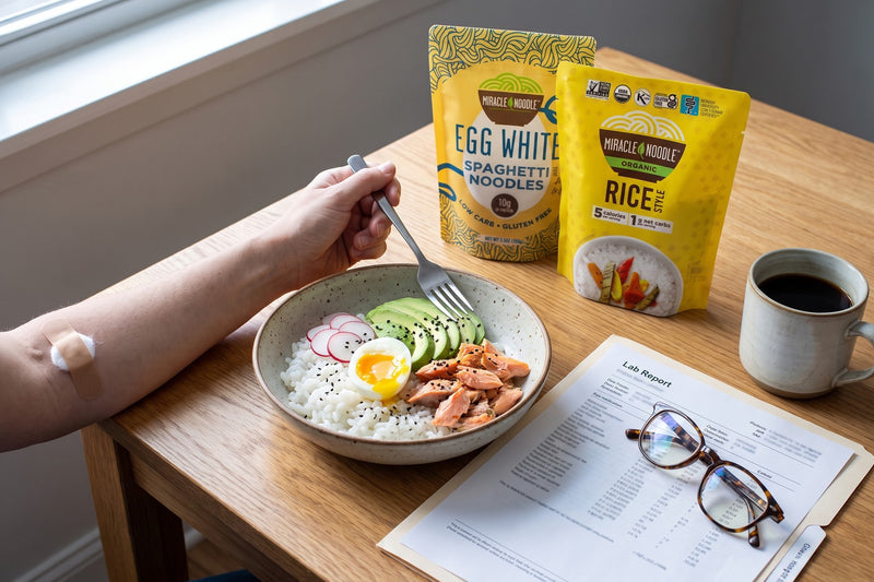 A forearm with a small bandage beside a rice bowl with a soft egg, trout and avocado, a lab report and glasses, with Miracle Noodle Egg White Spaghetti and Rice pouches