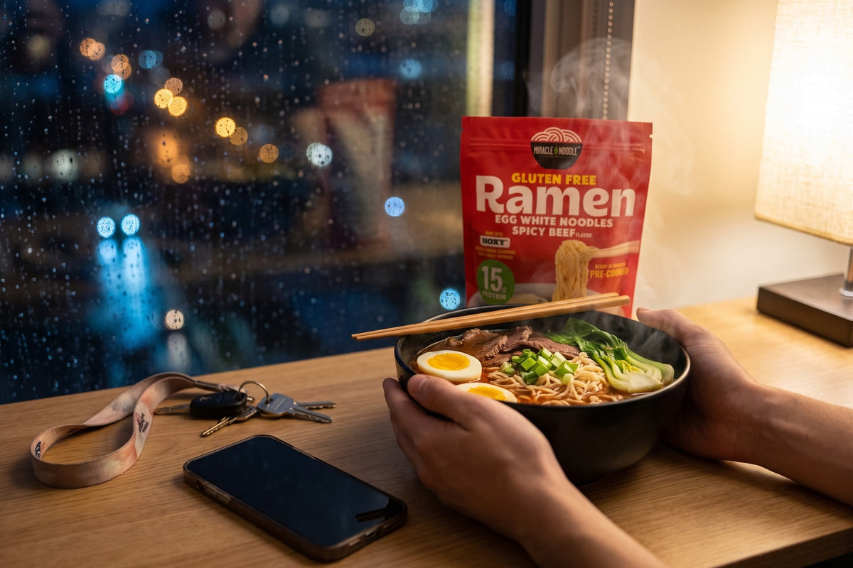 Two hands around a steaming bowl of spicy beef ramen by a rain-streaked window at night, with a Miracle Noodle Ramen pouch behind