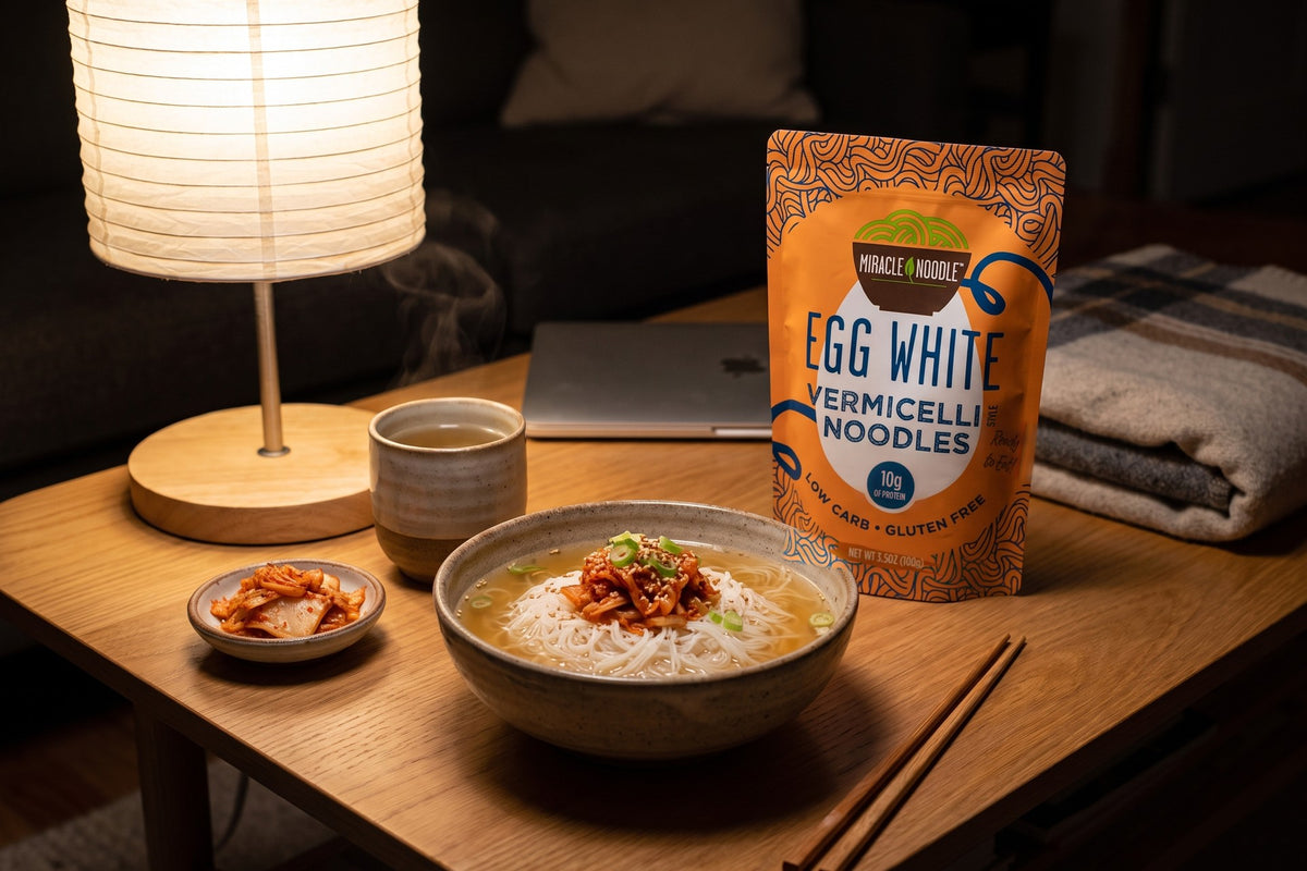 A bowl of egg white vermicelli in broth topped with kimchi under a paper lamp, with tea, a closed laptop and a Miracle Noodle Egg White Vermicelli pouch