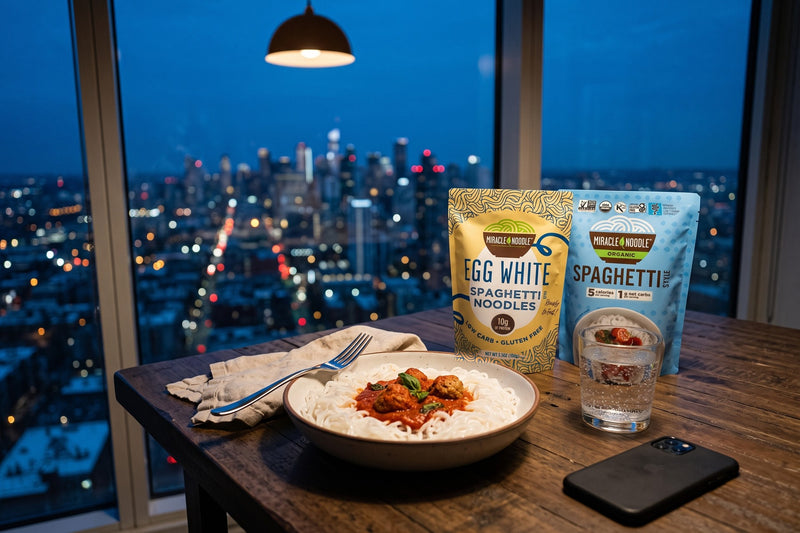 A bowl of white spaghetti with tomato sauce and meatballs on a table by a window over a city at dusk, with Miracle Noodle Egg White Spaghetti and konjac Spaghetti pouches