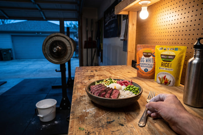 A bowl of konjac rice with sliced seared steak, edamame and pickled onion on a garage-gym workbench, a chalked hand reaching for a fork, with Miracle Noodle Egg White Vermicelli and Rice pouches