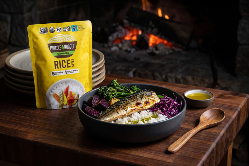 A bowl of konjac rice with grilled mackerel, broccolini, beets and red cabbage in front of low embers, with a Miracle Noodle Rice pouch