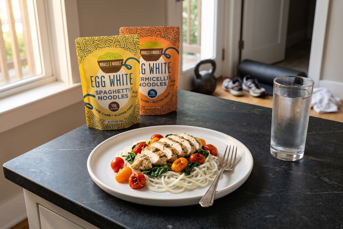 A plate of egg white spaghetti with grilled chicken, spinach and tomatoes on a dark counter, a kettlebell by the door, with Miracle Noodle Egg White Spaghetti and Vermicelli pouches