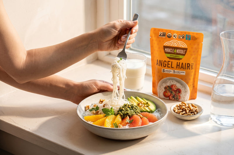 Hands lifting a forkful of konjac angel hair from a bowl with citrus, avocado, walnuts and pesto in morning light, with a Miracle Noodle Angel Hair pouch