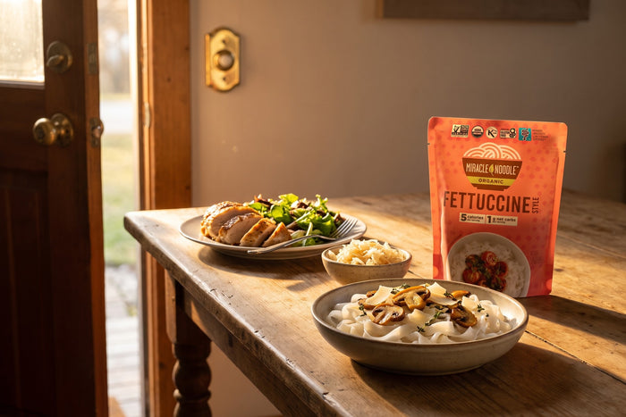 Roast chicken with salad, a small bowl of sauerkraut and a bowl of konjac fettuccine with mushrooms on a table by an open front door, with a Miracle Noodle Fettuccine pouch