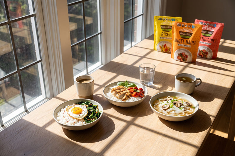Three bowls of konjac rice, angel hair and fettuccine in a row on a sunlit table crossed by window shadows, with Miracle Noodle Rice, Angel Hair and Fettuccine pouches