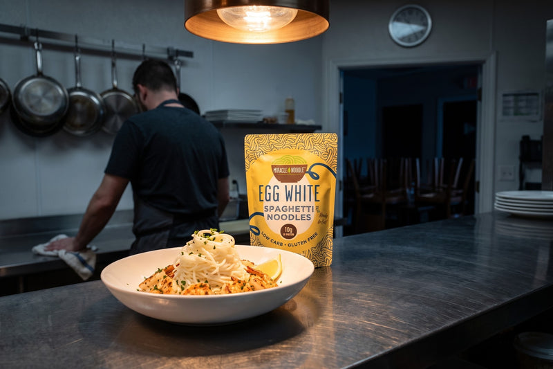 A bowl of egg white spaghetti with garlic shrimp on the steel pass of a restaurant kitchen being closed for the night, with a Miracle Noodle Egg White Spaghetti pouch